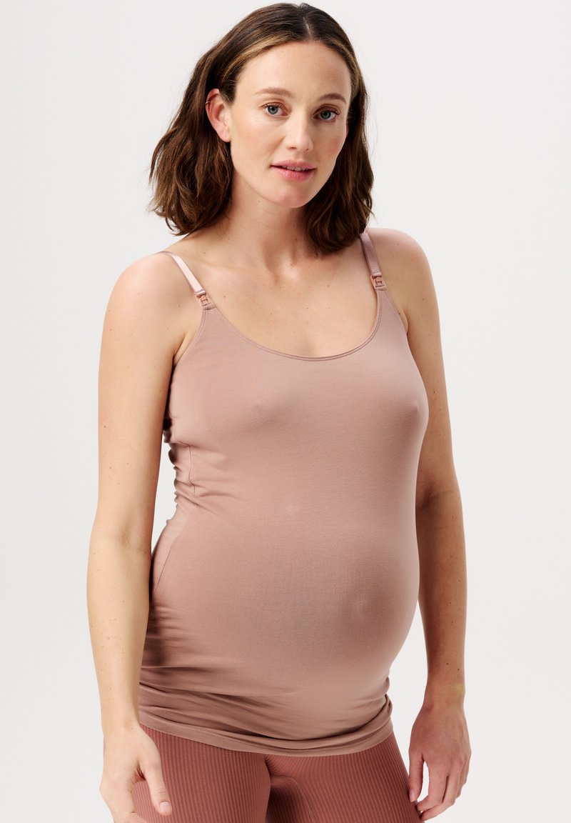 Gravid kvinde iført en tætsiddende lyserød tanktop og ribbede mauve leggings, stående foran en ensfarvet hvid baggrund.