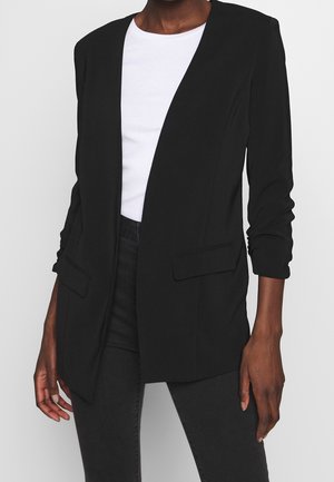 Blazer negro abierto al frente con bolsillos de solapa, usado sobre una camiseta blanca y pantalones negros, mostrado en una persona de pie desde el cuello hasta los muslos.