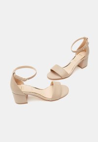Sandalias de tacón cuadrado beige con tiras en el tobillo, banda frontal ancha, textura similar al cuero suave y acabado mate. Presenta un cierre con hebilla dorada.