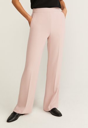 Kvinde iført lyserøde vidde bukser og sorte loafers, hænderne i lommerne, stående mod ensfarvet baggrund.