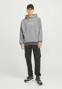 Sweatshirt à capuche gris avec logo sur le devant, pantalon cargo noir avec poches et chaussures de sport noires. Modèle portant une casquette noire.