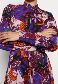 Mönstrad klänning med flerfärgade blommor i lila och svart, med en hög volangkrage, långa ärmar och smockade manschetter. Nyckelh ålsdetalj vid halsen.