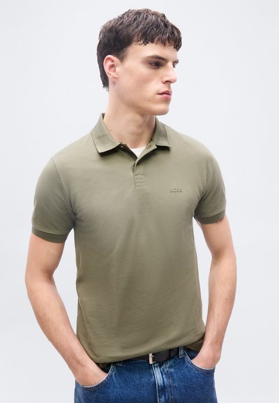 Jeune homme debout les mains dans les poches, portant un polo vert olive et un jean bleu, regardant sur le côté sur un fond uni.