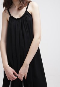 Black sleeveless dress with gathered fabric and a textured neckline, featuring a loose fit and subtle draping. Silky material with a smooth surface.