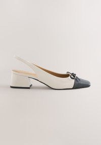 Chaussure slingback blanche à talon bloc avec bout noir pointu et petit nœud sur fond uni clair.