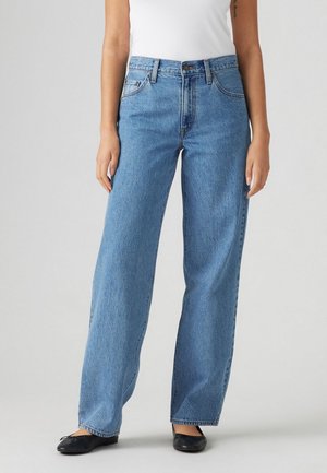 Femme portant un jean bleu taille haute droit, un top blanc sans manches rentré et des ballerines noires, debout devant un fond uni.