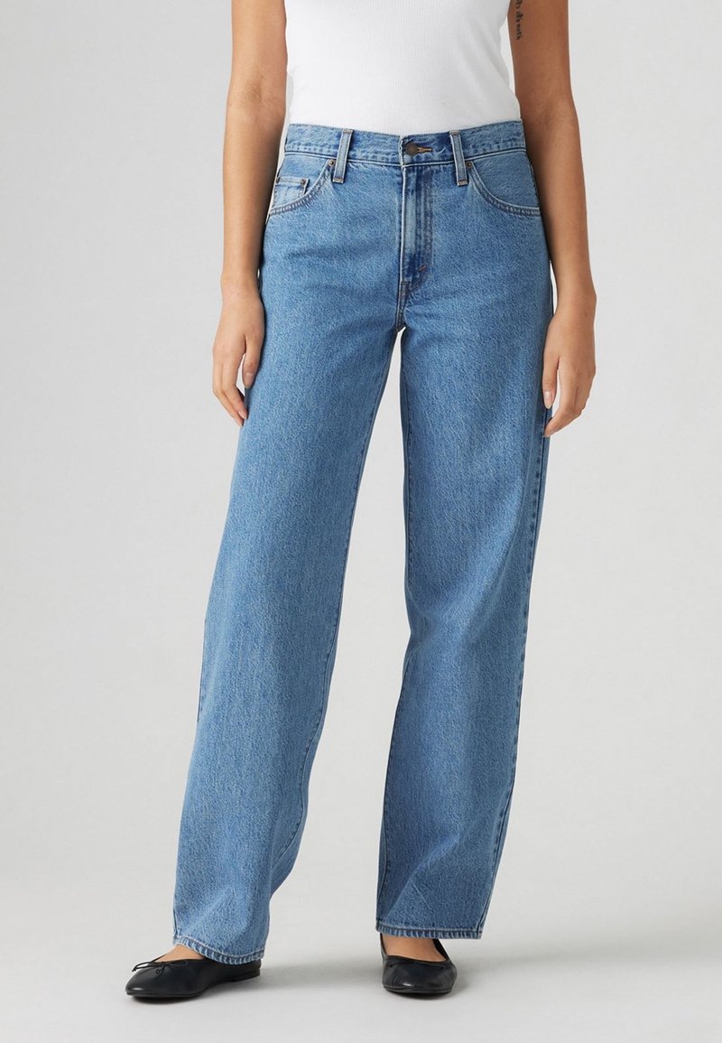 Femme portant un jean bleu taille haute droit, un top blanc sans manches rentré et des ballerines noires, debout devant un fond uni.