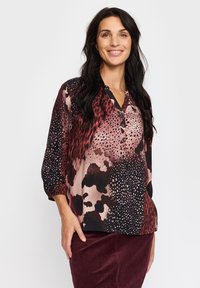 Trykt bluse med V-hals, lange ermer og knappedetaljer. Har en blanding av dyremønstre og prikkemønstre i dype røde og svarte farger.