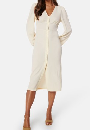 Mujer con un vestido midi color crema, de manga larga con botones, escote en V y tejido texturizado, combinado con tacones abiertos en tono tostado.