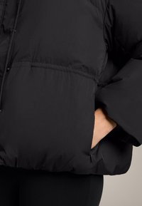 Čierna prešívaná bunda s jemným, textúrovaným povrchom. Má priestranný strih, elastické manžety a bočnú kapsu so zipsom.