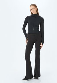 Chaqueta deportiva negra con cremallera, de manga larga y cintura ajustada, combinada con leggins negros acampanados. Textura suave y diseño minimalista.