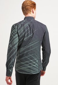 Uomo con capelli rossi corti che indossa una camicia a maniche lunghe scura con un motivo a strisce diagonali astratte verdi, girato di spalle rispetto alla fotocamera.