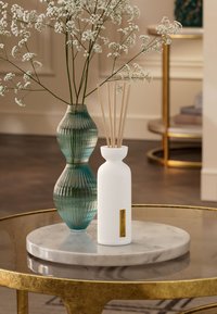Hvit ribbet diffuser med lyse trebunter ved siden av en teksturert turkis glassvase som holder hvite blomster på et marmorbord.