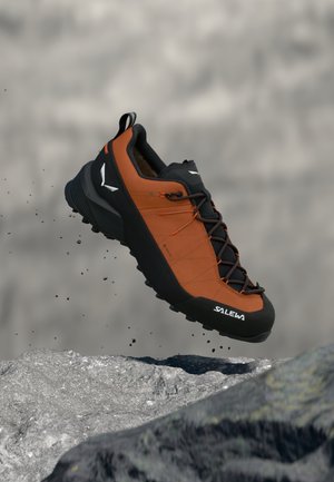 Zapatilla de trail naranja y negra con suela de goma, parte superior texturizada y puntera reforzada. Cuenta con sistema de cordones y logotipo en el lateral.