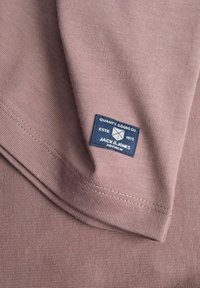 Material mov pliat cu o etichetă dreptunghiulară albastră mică cu inscripția „Quality Goods Co. Jack & Jones Estd 1975” cusută aproape de margine.
