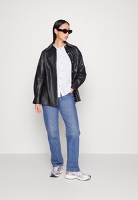 Casaco de couro preto, camisa branca com botões, jeans azuis e ténis prateados. Pequenos óculos de sol pretos completam o visual.