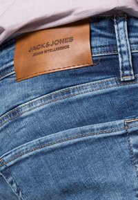 Jeans azules de mezclilla con un parche de cuero marrón de la marca etiquetado "JACK&JONES", con costuras visibles y detalles de textura en la tela.