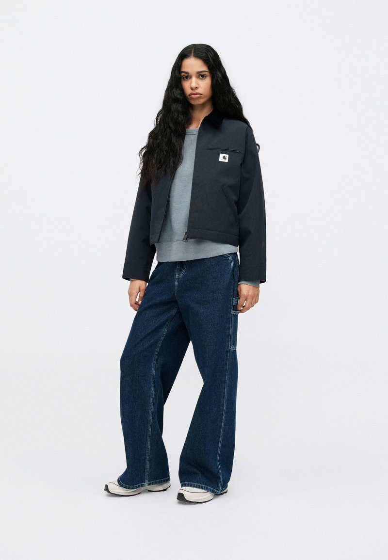 Mørkeblå cropped jakke med lynlås, grå sweatshirt under, og brede blå jeans. Funktioner inkluderer en frontlomme og sneakers.