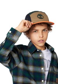 Il cappello presenta un pannello frontale marrone, lati blu navy e verde, visiera arancione e logo. Indossato con una camicia a quadri verde e blu navy con bottoni.