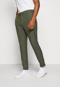 Olivgröna skräddarsydda byxor i ett glatt tyg, med flat front, bälteshällor och en enda knappstängning, tillsammans med vita sneakers.