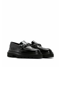Mocasines de cuero negro con un acabado brillante, que presentan una cadena decorativa en la parte superior y una suela de goma gruesa y texturizada.