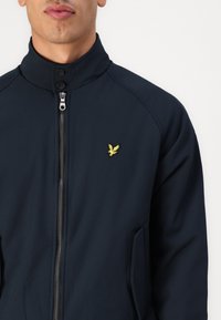Giacca zip-up blu navy con colletto alto, caratterizzata da un piccolo logo di un uccello giallo sul petto e tasche laterali; texture del tessuto liscia.