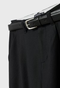 Pantalon noir sur mesure avec une ceinture en cuir noir présentant une boucle rectangulaire argentée, montrant partiellement l'étiquette à la taille.