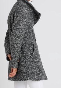 Abrigo gris texturizado con un amplio collar, bolsillos laterales y un diseño holgado, que presenta una mezcla de hilos oscuros y claros.