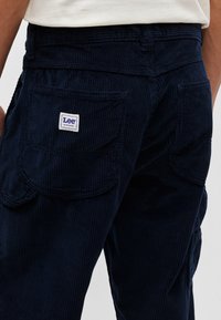 Pantaloni din corduroy navy, cu un design ajustat, buzunare cusute vizibil și un eticheta pătrată Lee pe spate. Material texturat cu caneluri.