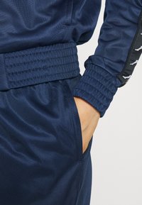 Main droite dans la poche d'un survêtement bleu avec taille et poignets élastiques côtelés, avec une bande noire et des logos blancs sur la manche.