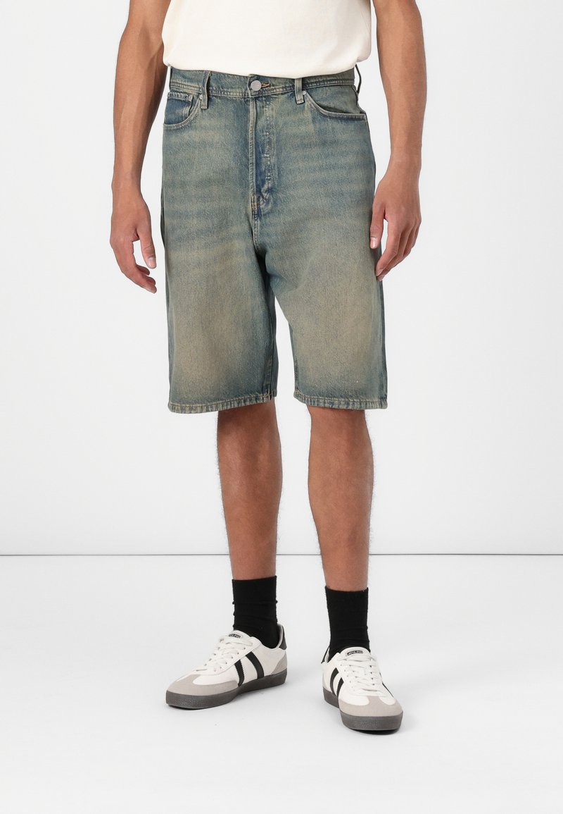Shorts de mezclilla, de color azul claro con desgastado, hasta la rodilla, de corte clásico. Combinados con zapatillas gris y blancas, calcetines negros. Diseño sencillo, cinco bolsillos.