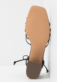 Vue de la semelle d'une sandale noire à lanières avec un bout carré et un patch en caoutchouc texturé sur fond blanc.