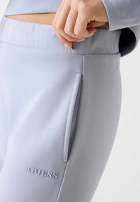 Jasnoszare spodnie dresowe wykonane z miękkiego materiału, z boczną kieszenią i haftowanym logo "GUESS" jako detalem. Gładka faktura i luźny krój.