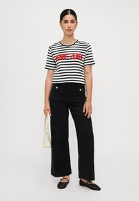 Randig svart och vit T-shirt med röd text "AMOUR", ihop med svarta vida byxor. Modellen håller en vit handväska. Svarta platta skor på fötterna.