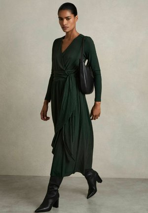 Abito lungo di un verde scuro con un design avvolgente e una texture plissettata. Abbinato a stivaletti neri con punta appuntita e una elegante borsa nera.