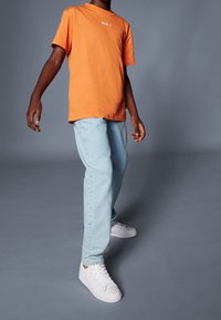 Camiseta de algodón naranja con un gráfico de texto blanco, combinada con jeans de mezclilla azul claro y zapatillas blancas con detalles en dorado.