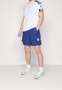 Maglietta atletica bianca con strisce nere sulle spalle, abbinata a pantaloni shorts blu con tasche laterali e una patch logo azzurro chiaro.