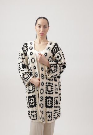 Kvinde med mørkt hår i en lav hestehale iført en sort og hvid hæklecardigan med geometriske mønstre over en hvid top og beige bukser.