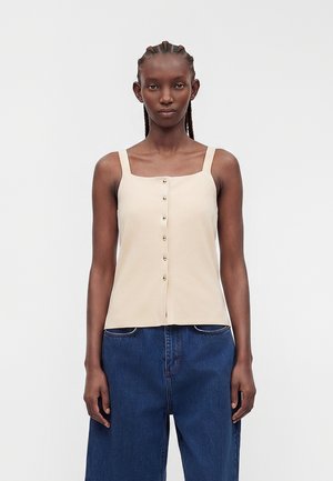 Kvinde med flettet hår iført en beige ærmeløs top med knapper foran og løstsiddende blå jeans, der står mod en ensfarvet hvid baggrund.