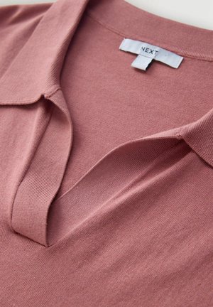 Putekļainas rozes krāsas polo krekls ar maigu faktūru, kurā ir izšūts apkakle un piekariņš ar uzrakstu "NEXT". Audums šķiet gluds un viegls.