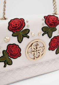Bílá prošívaná kabelka s červeně vyšitými růžemi, zelenými listy a zlatými akcenty. Obsahuje kruhové logo a dekorativní hroty.