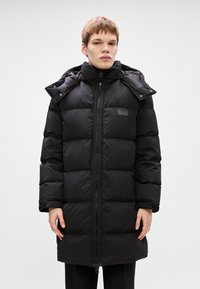 Black puffer coat featuring a high collar, zip closure, and snap buttons. It has a padded design with stitched panels and a logo patch.