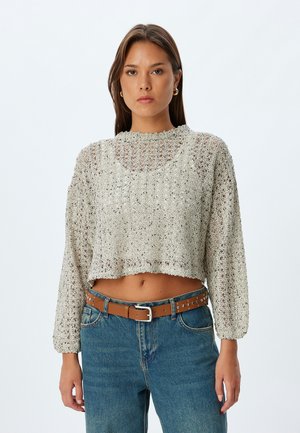 Maglione lavorato all'uncinetto di colore grigio chiaro, design cropped con maniche lunghe e colletto a coste, abbinato a jeans a vita alta e una cintura marrone.