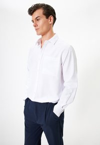Chemise blanche à manches longues avec une poche poitrine, devant à boutons et poignets, associée à un pantalon bleu marine foncé. Texture lisse.