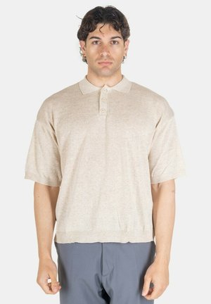Uomo con capelli scuri e ricci che indossa una polo beige a maniche corte e pantaloni grigi, in piedi davanti a uno sfondo bianco uniforme.
