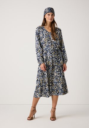 Marineblaues Blumenkleid mit langen Ärmeln, das mit gelben und weißen Blumen verziert ist, tailliert und kombiniert mit einem passenden Kopftuch und braunen Absätzen.