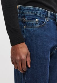 Dark blue denim jeans with a straight fit, five pockets, subtle whiskering, and copper stitching. Worn with a black sweater.