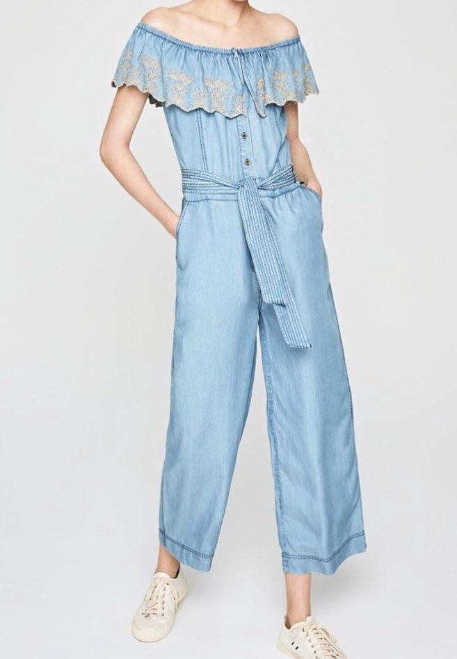 Combinaison bleu clair épaules dénudées avec col à volants brodé, ouverture boutonnée devant, ceinture à la taille, jambes larges et baskets blanches.