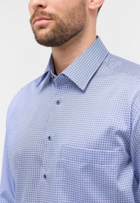 Camicia blu con un motivo geometrico, maniche lunghe, colletto con bottoni e tasca. Presenta bottoni azzurri chiari e una texture liscia.