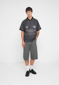 Svart kortermet genser med diamantmønster, har svart krage og hvit tekst. Sammen med grå knelengde shorts og svarte sko.